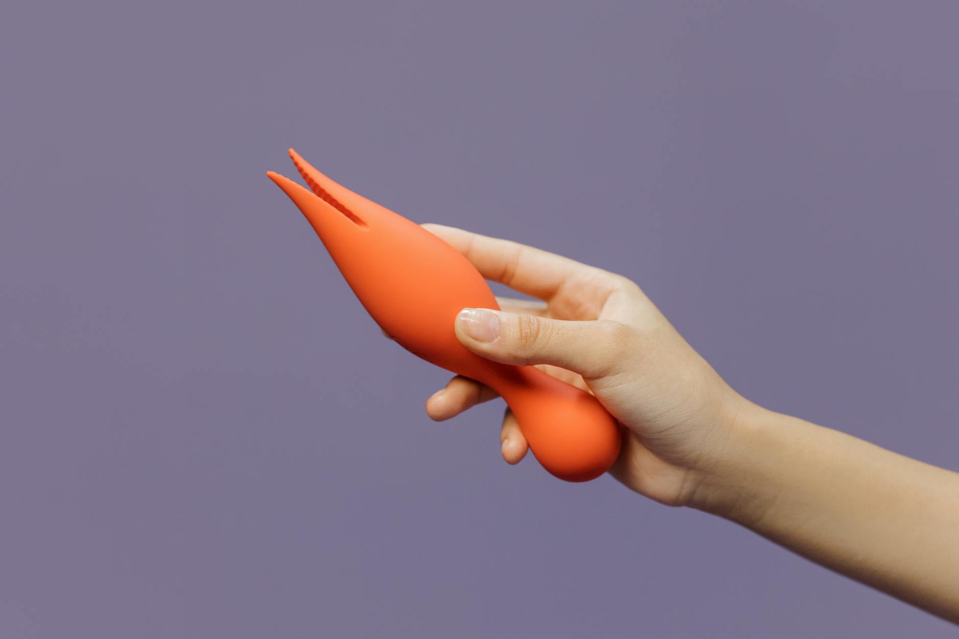 A hand holding a modern orange vibrator against a minimalistic purple backdrop