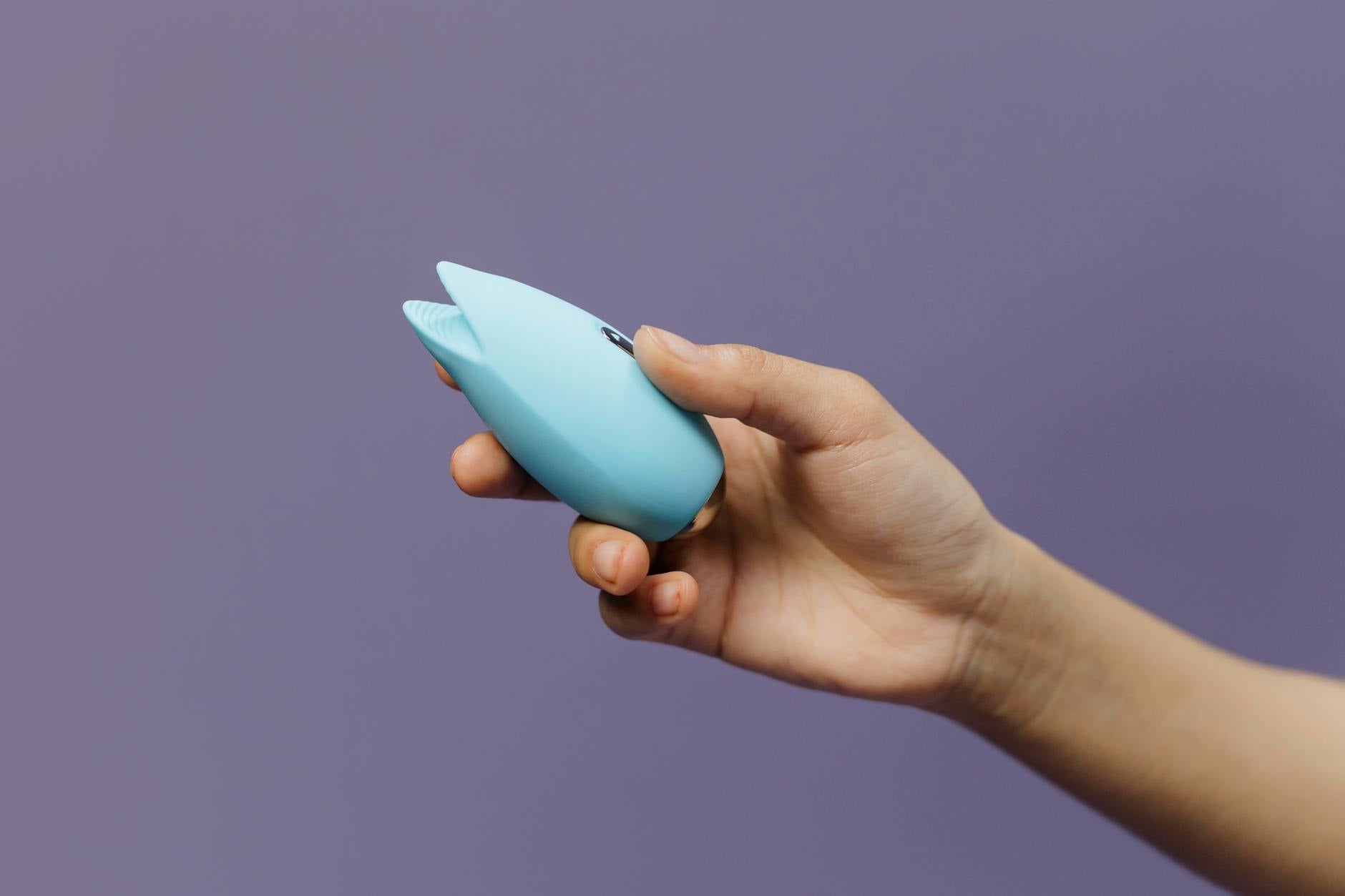 A blue silicone clitoral vibrator held in hand against a purple background, promoting reconnection and self-pleasure